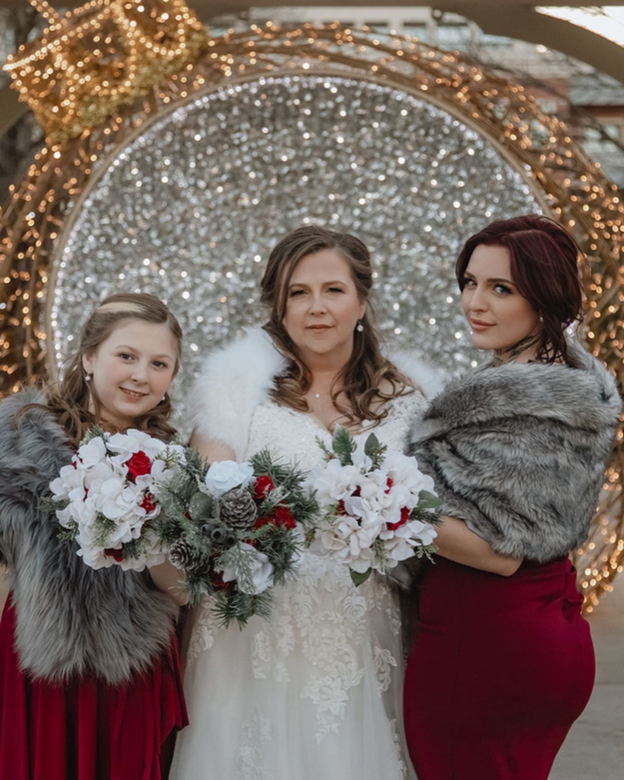 Bride flanked by her bridesmaids in faux fur wraps, holding winter bouquets before a sparkling arch
